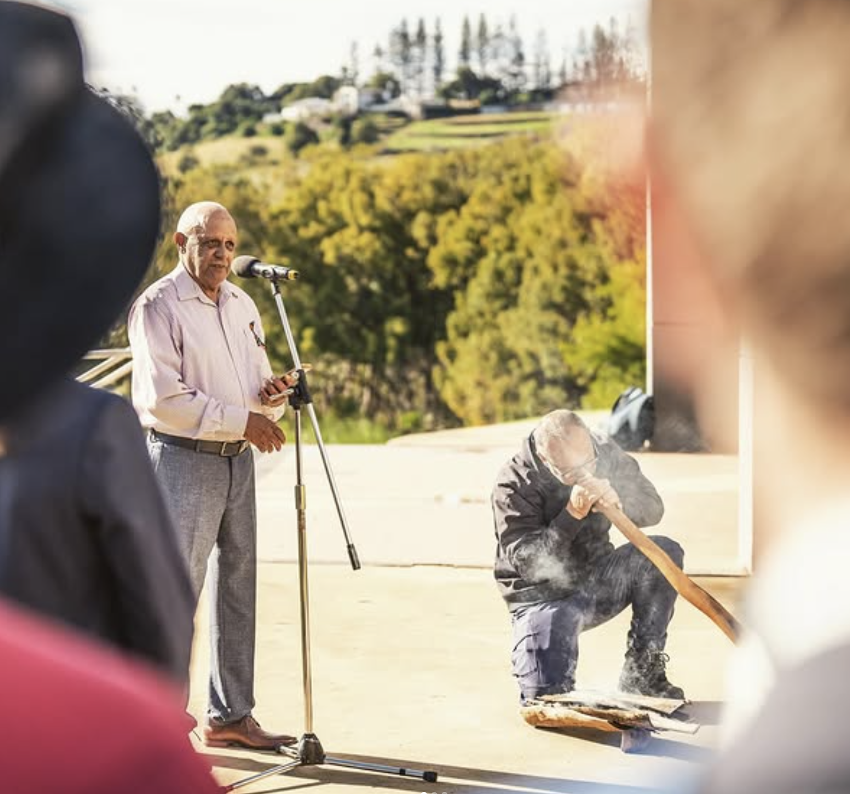 A man stands at a microphone, speaking to an audience, while Shannon plays a didgeridoo in the background.