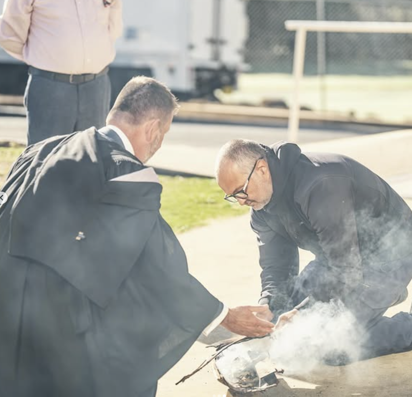 Two men in dark clothing preparing a fire pit on a sidewalk, with one man kneeling and the other standing nearby.